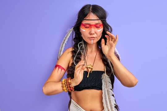 indian shamanic woman with warrior shaman make up isolated in studio on purple background, colourful paintings on her face and body, she is looking down shamaning