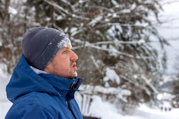 Portrait of a young man out in nature during snowy day in winter. Shot of a man wearing a winter coat standing in a snowy landscape. Photo of a young man with beard standing in the snowy mountains.