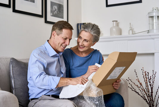 Happy old senior couple unpacking shipping delivery box sitting at home on sofa. Mature 50s grandparents customers unboxing parcel package receiving online shop order gift by postal shipment service.