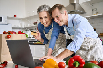 Happy senior old couple using laptop computer holding credit card ordering food delivery in grocery e store supermarket doing ecommerce shopping, buying online on website together at home in kitchen.