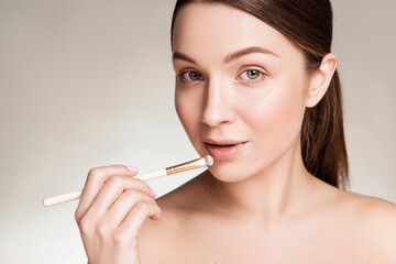 Beautiful brunette caucasian young woman prepare herself, applying lipstick on her lips with a brush. Clean, fresh, natural, flawless skin. Soft smile on her face. Close up on a neutral background