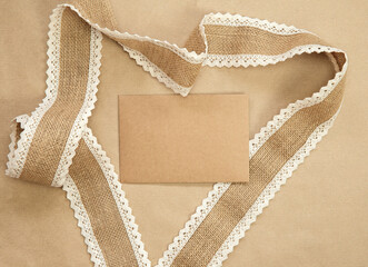 Flat Lay composition with blank brown plain greeting card surrounded with natural burlap ribbon on craft paper background texture, top view design, copy space natural colors