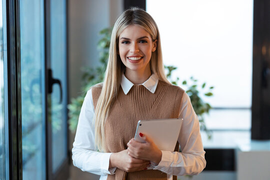 Pretty young businesswoman looking at camera while holding digital tablet standing in the office.