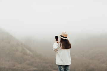 Fototapeta premium Woman is looking at foggy mountain and take photo on her phone. 