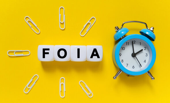 On a yellow background, a blue alarm clock, paper clips and white cubes on which the text is written - FOIA