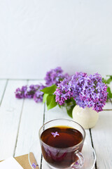 Delicate morning tea table setting with lilac flowers, a transparent cup of tea and a saucer and a white vase, white and craft note paper on a white wooden board. Mockup. 