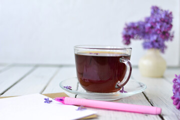 Delicate morning tea table setting with lilac flowers, a transparent cup of tea and a saucer and a white vase, white and craft paper for notes, and a pink pen on a white wooden table. Mockup. 