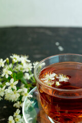 A transparent cup of teapot tea with a saucer and a white bird cherry flower on a black background. Copy space. Spring breakfast