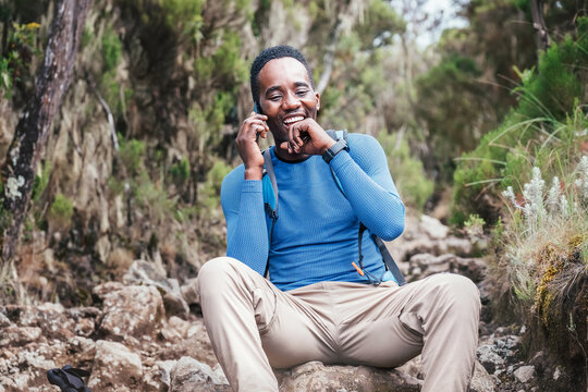 Young African ethnicity man talking via cellphone with somebody and cheerfully laughing as he having a hiking walk in the tropical forest. Happy people, network technology, or traveling concept.