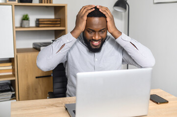 Young and handsome African-American businessman received exciting and positive news and cannot believe his eyes, looking at the laptop screen and holding his head in hands, luck man won a lottery