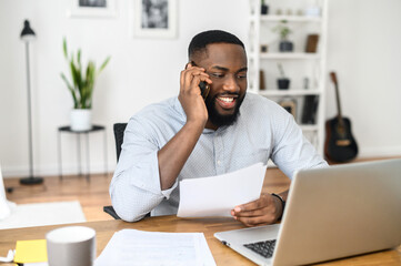 Fototapeta na wymiar Handsome African-American millennial businessman working in the office, sitting at the desk, holding a smartphone, solving business issues. Successful company member employee talking to a client