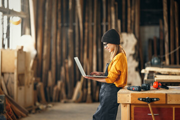 young female carpenter looks drawings on a laptop during a break in work in  workshop - obrazy, fototapety, plakaty