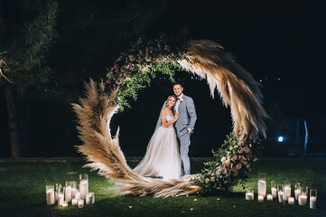 A young groom in a gray suit and a cute, smiling blonde bride embrace, standing on the green grass,...