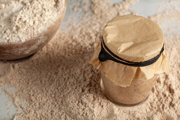 Homemade sourdough bread making ingredients featuring a bowl of flour and a sourdough starter culture in a glass cup covered with a brown paper on top. They all sit on flour covered kitchen countertop
