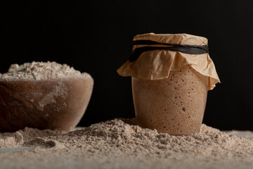Homemade sourdough bread making ingredients featuring a bowl of flour and a sourdough starter culture in a glass cup covered with a brown paper on top. They all sit on flour covered kitchen countertop