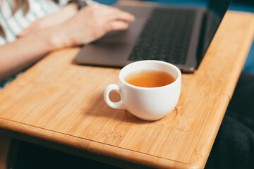 Close up photo of cup of tea on woman working on laptop in bed background.