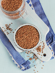 Organic uncooked scattered buckwheat grain in a bowl and glass jar on a light blue background. Healthy and diet food concept.
