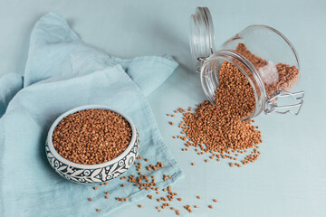 Bowl of dry raw buckwheat groats and glass jar on a light blue background. Cooking buckwheat...