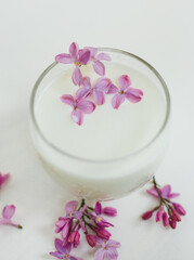 Obraz na płótnie Canvas A glass of milk and lilac flowers floating in it. White table. Simple still life.