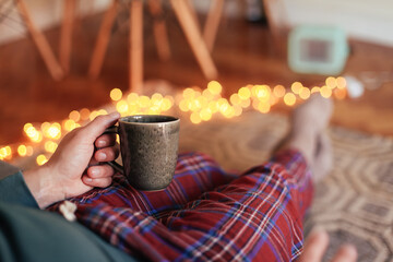 Man holding a cup of tea, wearing check pajamas, sitting at home in a cosy room
