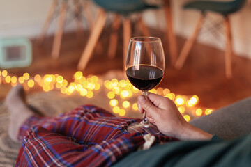 Man holding a glass of red wine, sitting on a couch in a cosy room, in red check pajamas