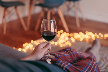A glass of red wine in hand, red check pajamas, cosy lights, a man sitting at home in the evening 
