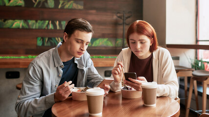 A couple of teenagers totally absorbed in using their phones, ignoring each other while sitting in a cafe together on a daytime