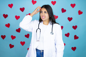 Young doctor woman wearing medical coat and stethoscope over blue background with red hearts confuse and wonder about question. Uncertain with doubt, thinking with hand on head