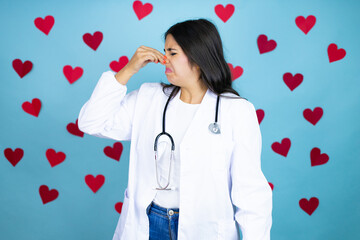 Young doctor woman wearing medical coat and stethoscope over blue background with red hearts smelling something stinky and disgusting, intolerable smell, holding breath with fingers on nose
