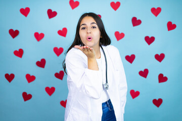 Young doctor woman wearing medical coat and stethoscope over blue background with red hearts looking at the camera blowing a kiss with hand on air being lovely and sexy. Love expression.