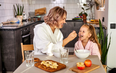 mother feeding her daughter by spoon, eating porridge, fresh fruits in the kitchen