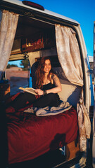 Obraz na płótnie Canvas Chica pelirroja en una furgoneta camper camperizada en una zona costera de playa al atardecer leyendo y escribiendo con estilo libre hippie