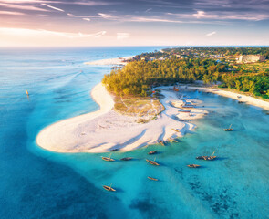 Aerial view of the fishing boats on tropical sea coast with sandy beach at sunset. Summer travel in Zanzibar, Africa. Top view of boats, yachts, green palm trees, clear blue water, colorful sky