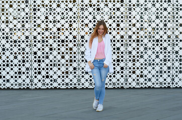 a girl in jeans and a white jacket on the street near a white wall