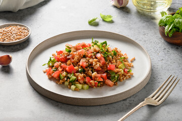 Useful vegan salad of whole grain cereal spelt with vegetables, tomato, cucumber, greens on grey. Close up.