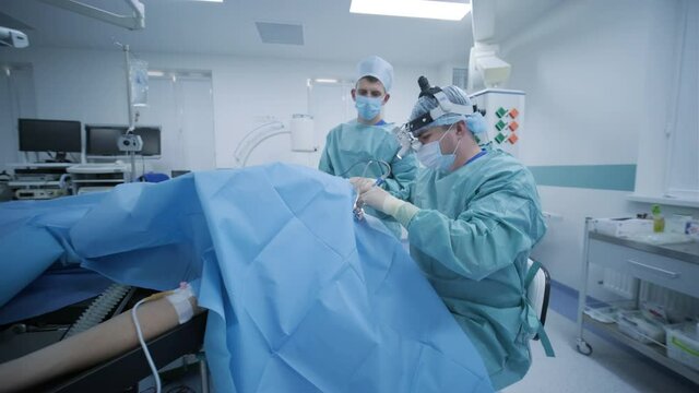 Operating room, patient is under deep anesthesia, surgeon with assistant uses latest devices, cold plasma method, cuts swelling in throat, doctor works with medical headlamp, surgical loupes