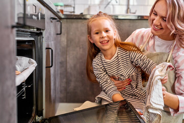 Fototapeta na wymiar daughter and mother taking out tray of baked cookies from oven in kitchen, family at home, baking and cooking concept