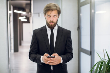 Front view of purposeful young businessman in elegant suit standing in modern office hallway and messaging on smartphone, while looking at camera. Close up portrait