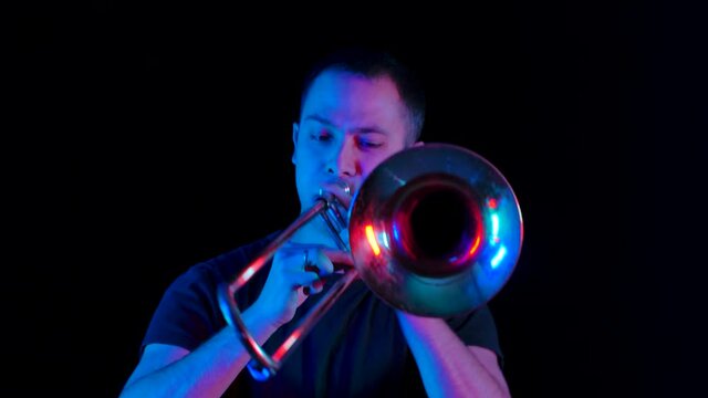 Front view of a musician playing the trombone in the studio against a black background in neon light. The thrombist performs music in the style of ska punk on a wind instrument. Close up. Slow motion.