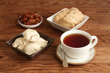 Meringue cookies with hazelnuts, almond cookies in a square bowl and pecan nuts in a small bowl, and a cup of black tea on a wooden table. Closeup