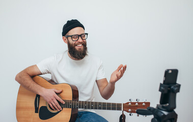 A young stylish guy with a beard wearing casual or music teacher playing guitar in front of smartphone camera. Online guitar training or vocal lesson