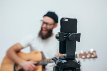 A young stylish guy with a beard wearing casual or music teacher playing guitar in front of smartphone camera. Online guitar training or vocal lesson