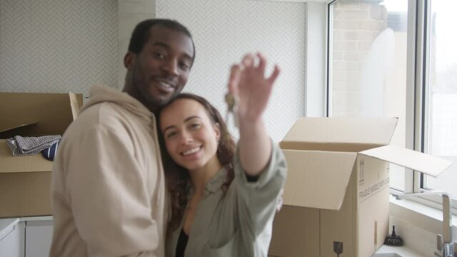 Portrait of young mixed ethnicity couple holding keys to new home standing in kitchen hugging as they unpack removal boxes together - shot in slow motion