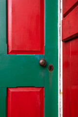 The exterior of a vintage red wooden shingled wall of a building with a bright red and vibrant green criss cross door. There are a rusty doorknob and keyhole in the door. The view is of the corner.