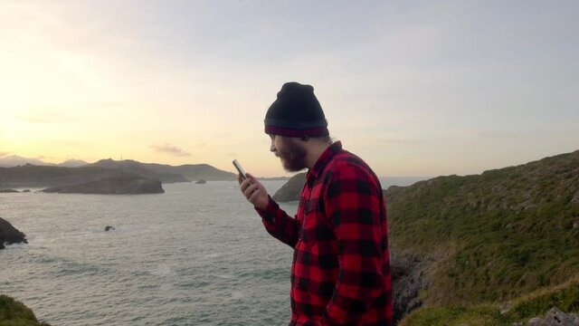bearded man in red plaid shirt using your smartphone with hands-free activated. pissed off person calling on the phone with the sea in the background. communications