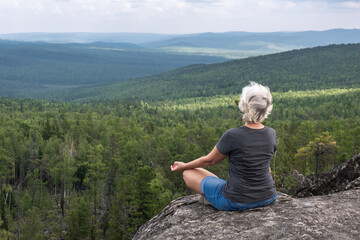 Naklejka premium A middle aged woman practices yoga and meditation on a mountain top with a stunning view of a forested, hilly valley.