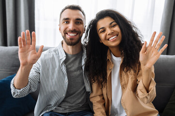 Multiracial couple in love, caucasian guy and african american girl, sitting on a sofa talk on video call with friends or family greeting, smiling and waving hands