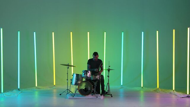 Expressive male musician plays drums in the studio against a background of multicolored neon tubes. A drummer with tattoos on his arms plays rock music with enthusiasm. Music concept. Slow motion.