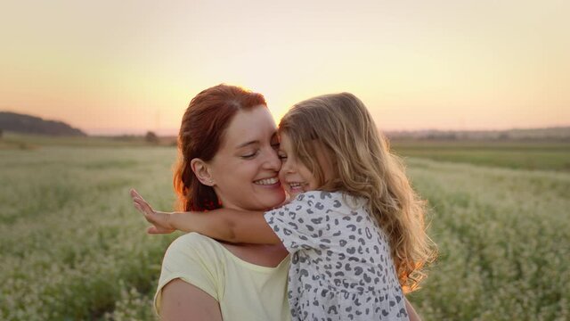 Young Mother Holding Her Daughter In Her Arms. They Are In The Middle Of The Field. They Are Both Smiling. Girl Kisses Mom.