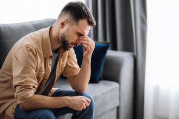 Tired guy sitting on the sofa at home, having a terrible headache. Exhausted caucasian male massage the eyes, feeling unhealthy, catch a cold or having flu, upset about illness